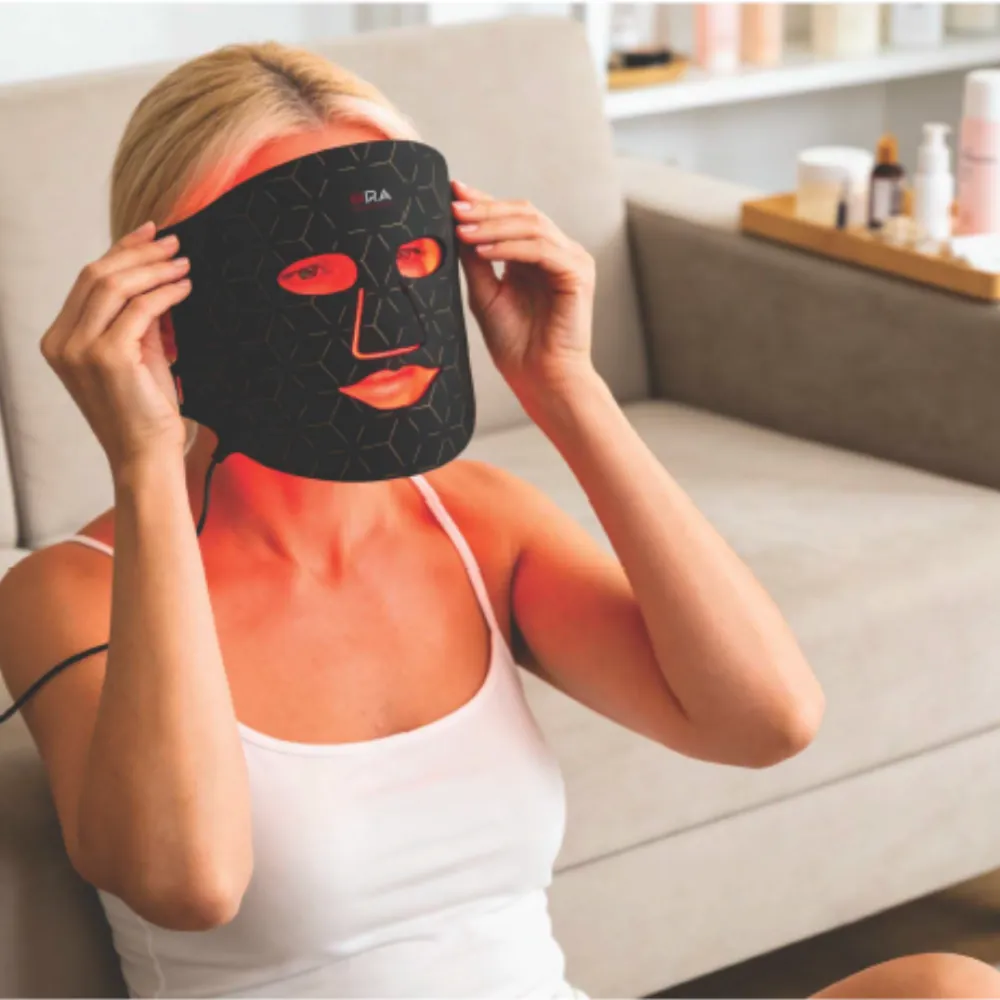 A woman sitting on a sofa putting on the ORA LED face mask, which glows red around the eyes and mouth
