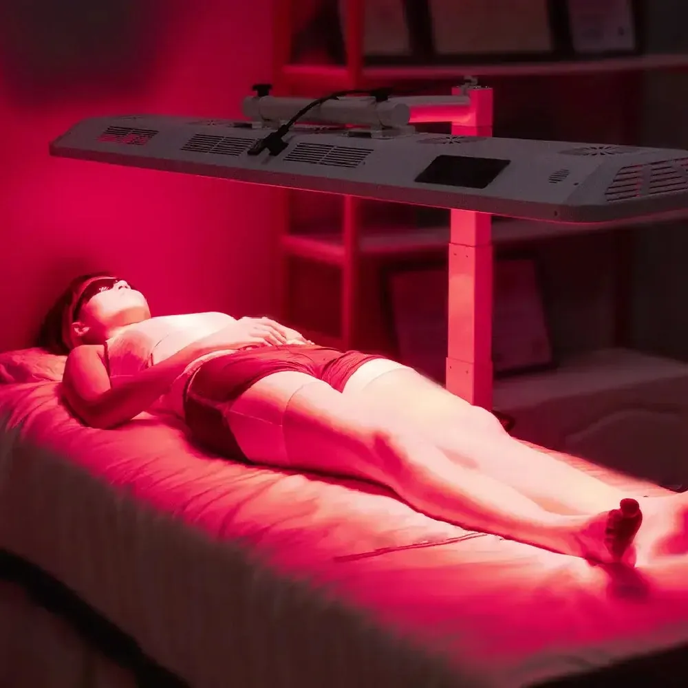 A woman resting on a treatment bed beneath a large overhead red light device