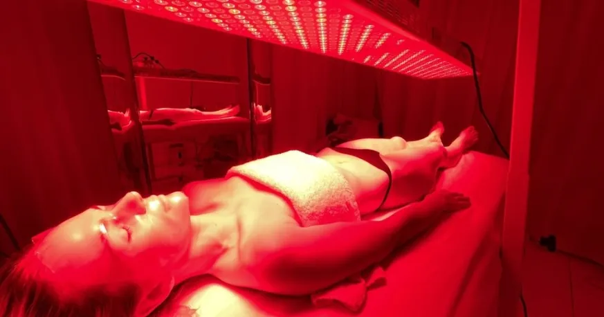 A woman lying on her back under a long overhead red light panel in a red-lit treatment room