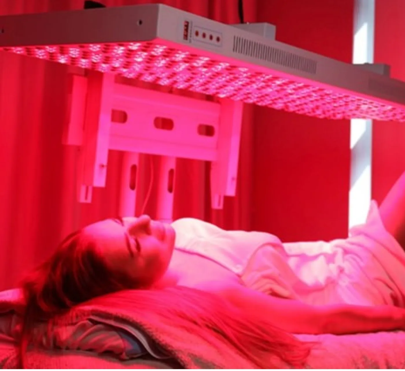 A woman relaxing on a treatment bed under a large overhead red light panel, with the room bathed in red light